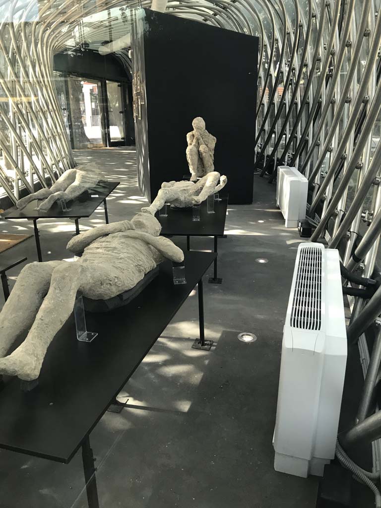 Pompeii. April 2019. General view of plaster-casts in the exhibition kiosk near Amphitheatre entrance.
Photo courtesy of Rick Bauer.
Left front, plaster-cast of victim 13, a man who was found in 1889 just outside the Stabian Gate.
He was described as lying on his back, wrapped in a cloak, and with his hands held as if protecting his chest.
His legs were slightly bent.