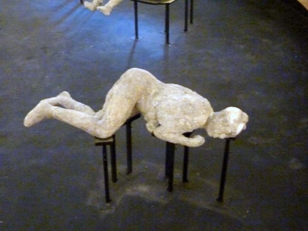 Victim 54. Cast of a victim made between 31st of July and 21st of September 1976.
Plaster cast of victim on exhibit in display in temporary pyramid in amphitheatre. September 2015.
Victim 54 is a female aged over 20 years.
The body was found outside Porta Nola, by the Tomb of M. Obellius Firmus, between the 31st of July and the 21st of September 1976.
The imprint of the clothes, perhaps a tunic and a cloak, rolled up between the thighs, on the abdomen and back is clearly visible; on the right shoulder the drapery is evident and on the left calf the imprint of the weave of a light fabric seems to be distinguishable; there is possibly the presence of a headdress and sandals, traces of which appear to be visible on the left foot.
On the 15th of September 1976, next to the left hand of the victim part of an iron ring with a gem incised with an image of the goddess Fortuna.
On the 31st July, before the cast was made, near the body were found a gold ring with an emerald, a silver ring with the head of a serpent, another gold ring with bezel bearing the engraving of a palmette or ear of corn, a small silver ring with a chain to which a phallic pendant must have been hung, a silver statuette of Isis Fortuna and a hoard of money consisting of four silver denarii and twenty bronze coins.
See Osanna, N., Capurso, A., e Masseroli, S. M., 2021. I Calchi di Pompei da Giuseppe Fiorelli ad oggi: Studi e Ricerche del PAP 46, p. 446-448, Calco n. 54.