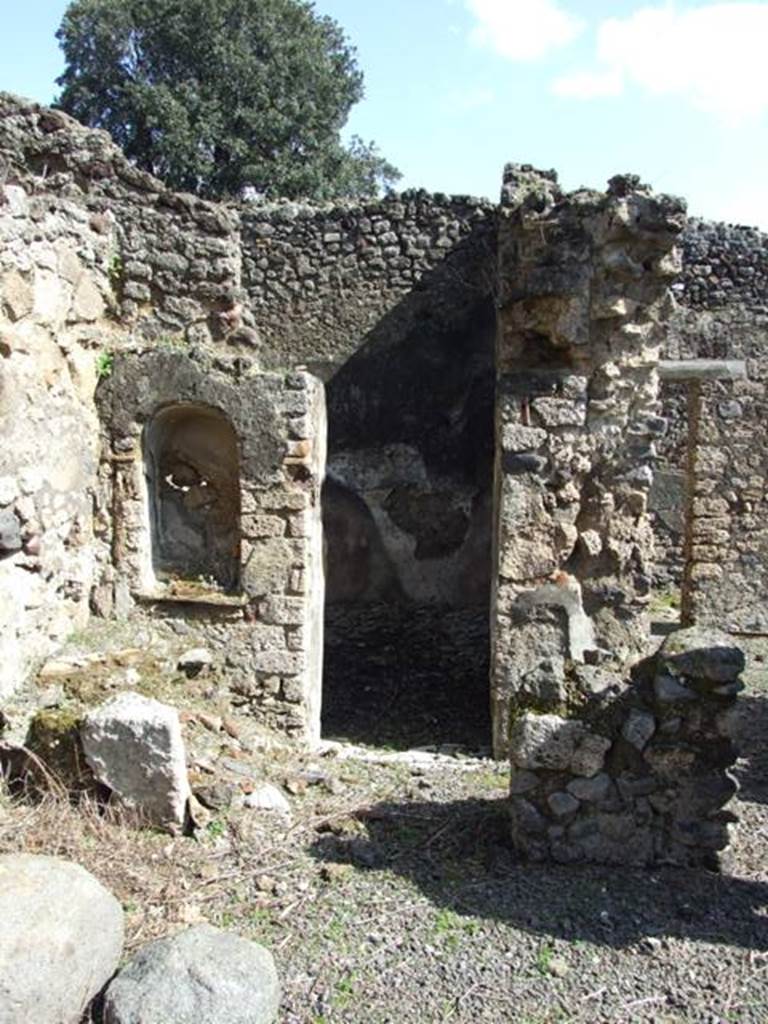 V.4.9 Pompeii. March 2009. Kitchen on east side of atrium. Looking east. According to Boyce, on the south-east corner of the impluvium was a square column (see remains on right of photo). This column closed off part of the east side of the impluvium to provide a small area as a kitchen. In the west side of this column, facing the impluvium, was a niche with slightly arched ceiling and projecting floor. Above and to the right of the niche was a yellow laurel wreath with pendent taeniae hung from a nail – all in stucco relief (see photo below). Within the niche were found – an iron knife with bronze handle, an iron hammer, and a bronze rosette. On the same side of the column, but higher, was another larger rectangular niche, probably for storage purposes.
See Boyce G. K., 1937. Corpus of the Lararia of Pompeii. Rome: MAAR 14. (p.41, no.124, Pl.4,5)
Hardly anything remains of the column today, and neither niche.
In the north-east corner of the kitchen stood a small hearth and above it in the east wall was a high arched niche (see above photo, on the left). Its walls were coated with white stucco. On its floor stood a masonry block, apparently for use as an altar, behind it a step was built against the rear wall. In the centre of the step was a mark left by an oval object which had stood there, probably a statuette. Another altar, terracotta and cylindrical, was found within the niche.
See Boyce G. K., 1937. Corpus of the Lararia of Pompeii. Rome: MAAR 14. (p.41, no.125, Pl.4,5)