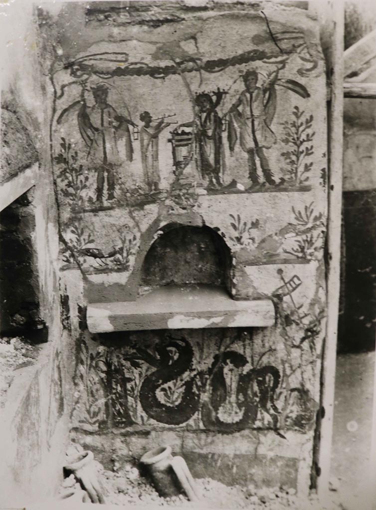 I.8.14 Pompeii. 1938.
Lararium painting and niche in north-east corner of courtyard 7, near to the kitchen (10), no longer conserved.
According to Giacobello, this is found in the north-east corner of viridarium 7 of I.8.14. Fröhlich had located this in insula I.8 but could not assign it to a specific house.
Above the niche is the painting showing two large Lares, a smaller togated Genius with a cornucopia and sacrificing on a round altar while on the other side of the altar a tibicen is playing the double flutes.
At the sides of the niche are two birds in shrubs.
Below the niche is a serpent approaching an altar.
See Giacobello, F., 2008. Larari Pompeiani: Iconografia e culto dei Lari in ambito domestico. Milano: LED Edizioni. (p.141, no.13).
Fröhlich describes a wall surface painted on a white background with an arched niche. In the uppermost zone, the sacrificing Genius stands on the right and Tibicen on the left at the round altar. The scene is flanked by two Lares, next to which a laurel bush stands on each edge of the picture. A garland is hung on the upper edge. The arched niche is recessed into the narrow, middle zone. On both sides of the niche, a peacock appears between small plants. In the lowest zone, a snake moves between marsh plants to the left towards a round altar. At the top right, a skewer with three pieces of meat is painted. In the right half, the remains of an older snake painting can be seen under the plaster, which may also include the adjacent zone with small plants, set off in color and by a plaster edge, to which a dark base is attached below. On the wall to the left, a pig's head is painted below and a ham above it. It is not known which phase these eating still lifes belong to. In any case, they suggest that the painting was located in a kitchen.
See Fröhlich, T., 1991. Lararien und Fassadenbilder in den Vesuvstädten. Mainz: von Zabern, See Fröhlich, T., 1991. Lararien und Fassadenbilder in den Vesuvstädten. Mainz: von Zabern, p. 254, L11, Taf. 26,2.