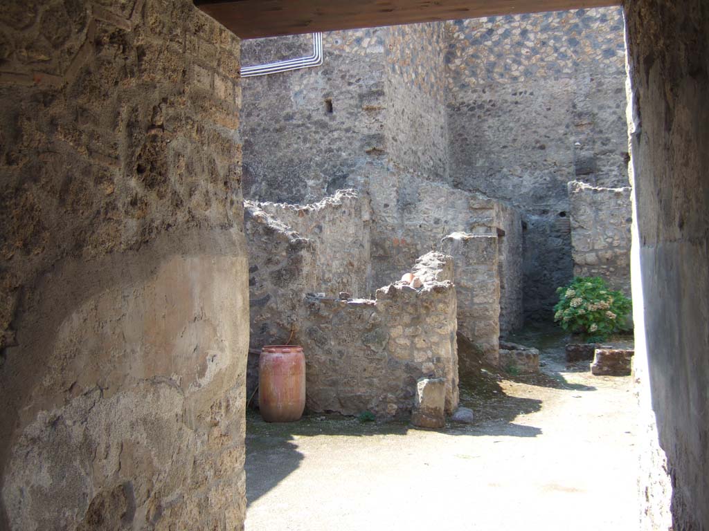 I.10.18 Pompeii. September 2005. Looking south-west from entrance fauces across atrium.
Right of centre is the area of room 6, looking ahead to cistern mouth in walkway No.8.
On the left at the end of the entrance corridor wall is the south-west side of the atrium.
Left of centre is room 7, the kitchen (room 9) is at the rear of room 7.
According to Boyce, in the south wall of the kitchen beside the hearth was a fragmentary painting in two zones.
It was on a white background outlined with red stripes.
In the upper zone stood the Genius, with a Lar.
In the lower zone, beneath garlands, was the tail of a black and yellow serpent.
Below the serpent were several kitchen articles, a hog’s head, a ham on a nail, and an eel on a spit.
See Notizie degli Scavi di Antichità, 1934, p. 344, and fig.38.
See Boyce G. K., 1937. Corpus of the Lararia of Pompeii. Rome: MAAR 14. (p.29, no.60)
Fröhlich described it was “mostly fallen from the wall, and only the body of the left Lar is quite well preserved.”
See Fröhlich, T., 1991. Lararien und Fassadenbilder in den Vesuvstädten. Mainz: von Zabern. (L18, Taf. 26, 3).