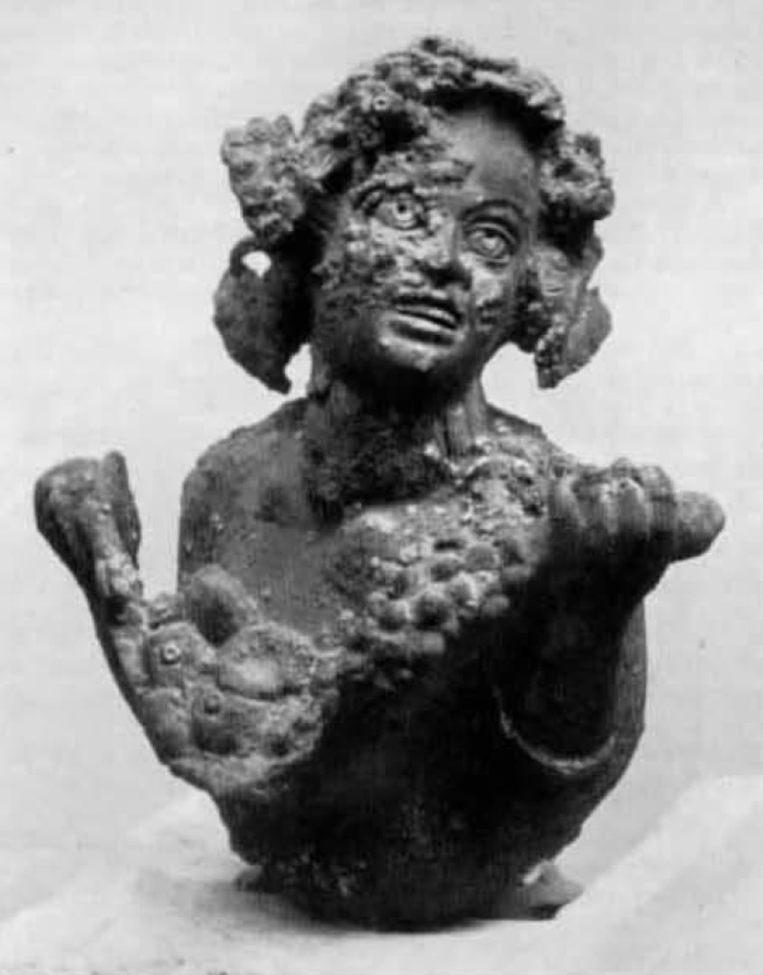 I.16.5 Pompeii. Small bronze bust of smiling satyr.
From the same room as the previous finding, and on the same day, the bronze bust had been found, depicting a satyr holding in his arms, a fleece (?) or a basket full of fruit.
The satyr turns up his laughing face, slightly leaning towards his left shoulder, the head of bunches of grapes, vine-leaves and branch of ivy make up a strange and lavish hairstyle.
The small bronze, which originally would have been an artistic part of furniture, probably of the bull (toro?) of a bed or a strong-box, has a maximum height of m.0.095 with a width of m.0.085.
Parco Archeologico di Pompeii. Inventory number 11872.
It was probably part of all the set of equipment, preserved/stored in the armarium
See Elia, O. Bacco Fanciullo e Dioniso Chtonio a Pompei. Bollettino d'Arte 1961, Fasc. I-II, (p.5, fig.7).