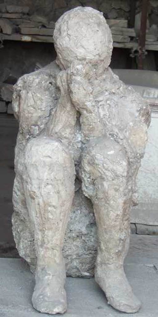 II.7.1 Pompeii. Palaestra.  Plaster cast of victim found near the latrine, perhaps found crouching on the ground with his back against the wall of the south portico.  Now in VII.7.29 Forum granary market.
Other victims found along the south portico (p.225) –  Skeleton of a horse and its driver.  The skeleton of the horse had been found almost leaning against the portico wall; his legs folded on the ground, his body a little contracted but still reassured in its anatomical composure; his head rested on the ash bank.  Less than a metre from the animal's head, the skeleton of the person to whom the horse belonged was found.
Extraordinary detail: eleven beads of glass-paste were collected around the bones of the beast's neck; a necklace as it is still used today in Campania as part of the decoration of the animal.
Found towards the eastern extremity from the southern portico, and from which plaster-casts could be made because they both lay in the high layer of ash. The one was a beautiful young man's body with agile legs knocked down on the portico floor at the first fall of the ashes.  The other, the most tragically posed, was found leaning against the wall, kneeling, crouching on the ground, in that crouching position that is found in Neolithic tombs, with his head folded forward, bent almost between his knees, for the better protection of himself with his hands and cloak against the exhalation of asphyxiating gases. He had shoes on his feet and wore the typical cloak of the peasant, the “cuculla”, whose flaps he held tight against his mouth.  And in removing the ash under his knees, imprints of overlapping fabrics were noticed, as if the unfortunate man had found comfort in his tragic pose by shielding his knees with the few garments he could arrange. (p.225-6)
For Maiuri’s “Scavo della “Grande Palestra” nel quartiere dell’Anfiteatro, -
see Notizie degli Scavi, Anno 1939, Fascicoli 7, 8, 9, on pages 165-238


