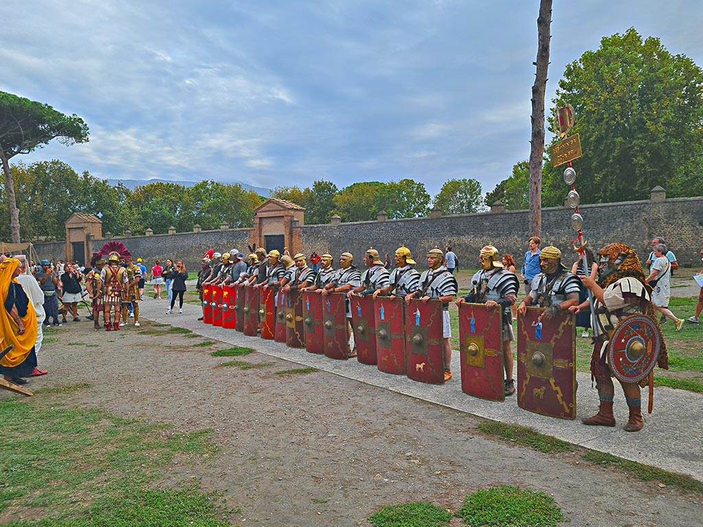 II.7.3 Pompeii, on left, II.7.4 in centre. September 2024. 
Looking south-west on Piazzale Anfiteatro towards Palaestra with legionaries on parade during “Ludi Pompeiani” event. 
Photo courtesy of Giuseppe Ciaramella.

