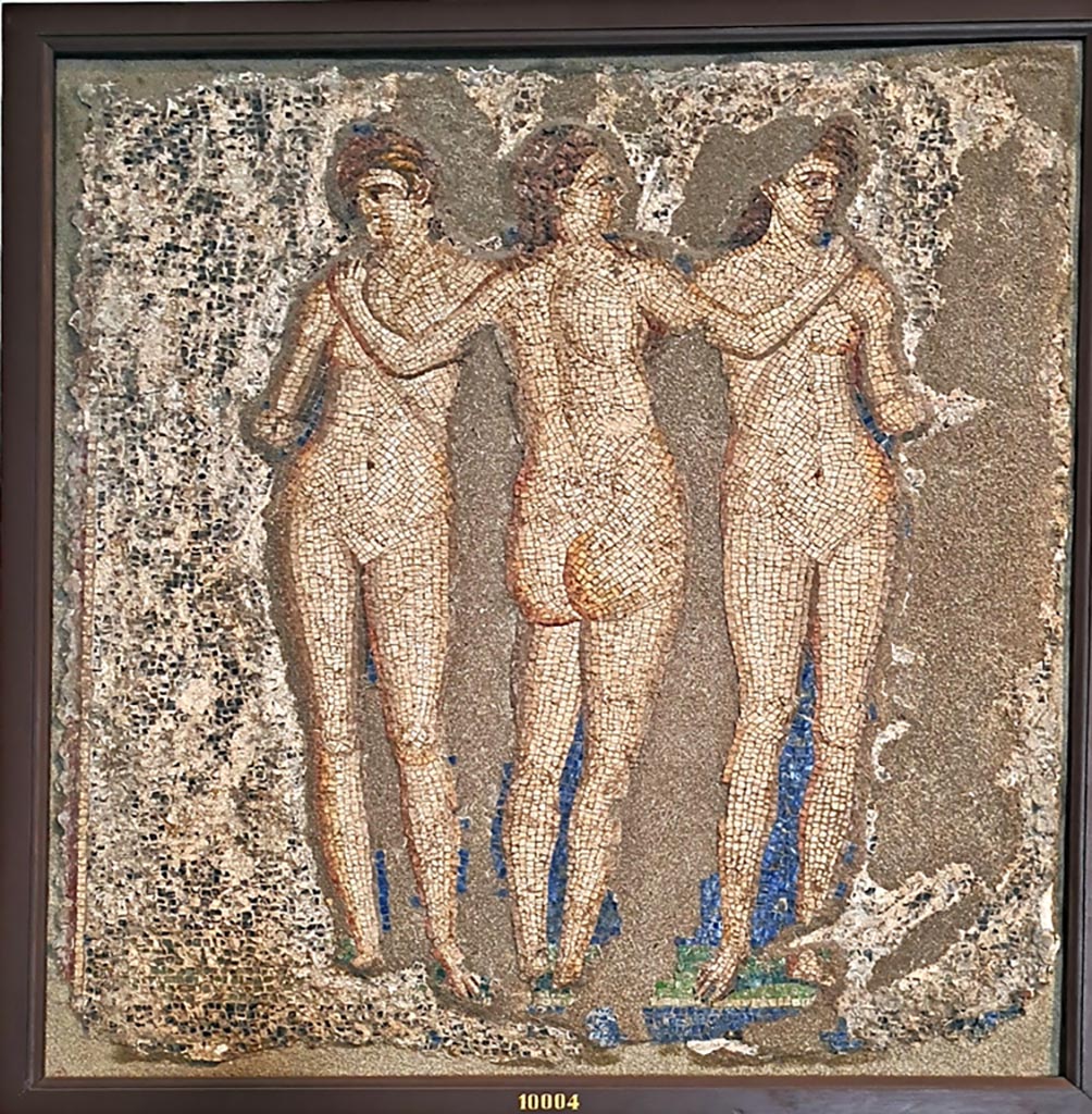 VI.7.23 Pompeii. 2024. Mosaic of the Three Graces from the garden wall.
Now in Naples Archaeological Museum. Inventory number 10004. Photo courtesy of Giuseppe Ciaramella
According to the information card –
The Three Graces. Opus vermiculatum wall panel made of limestone, glass paste and shells.
The three goddesses are sown dancing with their arms raised in the air in a gesture that gracefully unites them.
(Third quarter of the Ist century AD).
(Le tre Grazie. Pannello parietale in opus vermiculatum di calcare, pasta vitrea e conchiglia.
Le tre dee sono raffigurate, danzanti con le braccia sollevate all’altezza delle spalle in un gesto che le unisce con grazia.
Terzo quarto del I secolo d.C.)