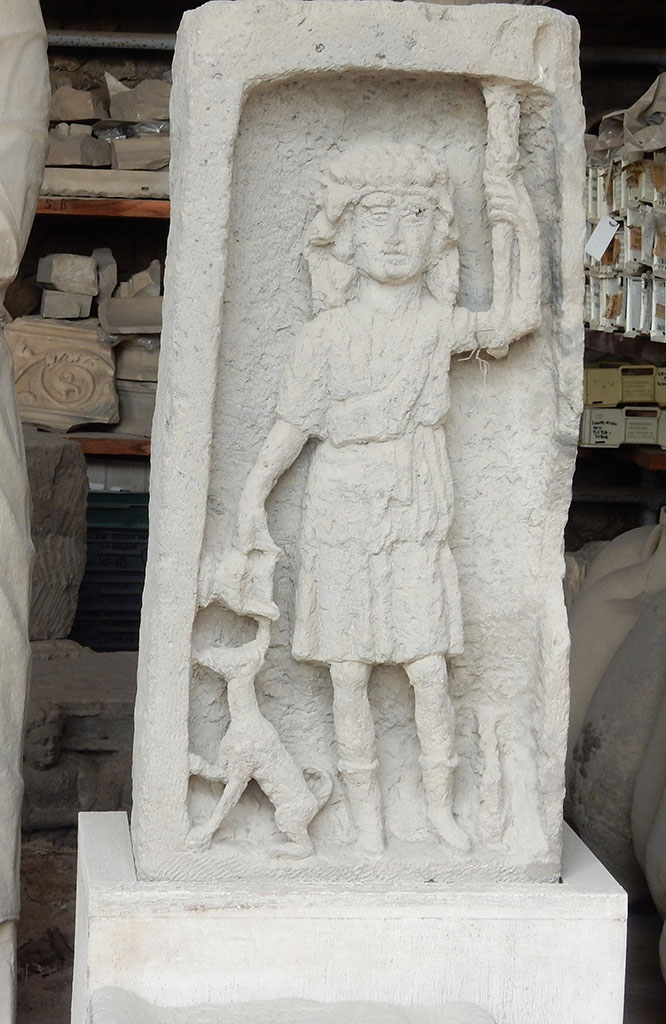 VII.7.29 Pompeii. May 2015. Bacchus stele in storage. Photo courtesy of Buzz Ferebee.
According to Kuivalainen, this a Bacchic votive stele of the Samnite era.
It depicts a front facing skinny youth with his weight on his right foot, pouring wine from a cantharus for an animal rather resembling a dog than a panther.
The middle part of the thyrsus is missing.
The width of the frame varies, being thinner on the right hand side.
It is cut deep into a rectangular upright tufa block and the workmanship is rather coarse.
The excavation details are not known.
PAP inventory number 8660.
See Kuivalainen, I., 2021. The Portrayal of Pompeian Bacchus. Commentationes Humanarum Litterarum 140. Helsinki: Finnish Society of Sciences and Letters, p. 207-8, H2.