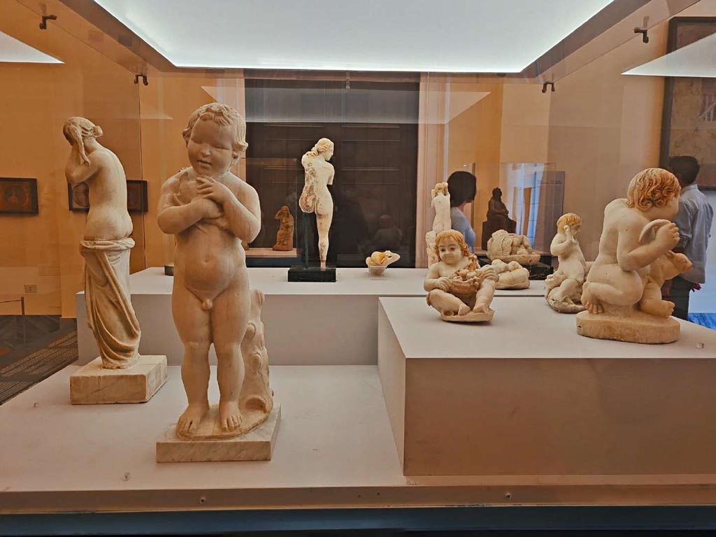 VII.12.23 Pompeii. October 2023. Group of statuettes, on right, found in the garden area.
On the left is a marble statuette of a child caressing a dove, from vesuvian area, but location unknown, (inv. 6114).
Photo courtesy of Giuseppe Ciaramella.
On display in “L’altra MANN” exhibition, October 2023, at Naples Archaeological Museum.