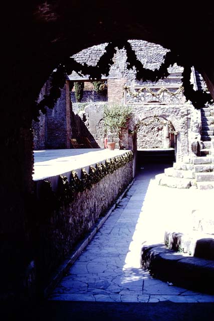 VIII.7.19 Pompeii. May 2006. East side with arched entrance. Above the entrances on either side were the rectangular areas which had the seats reserved for the honoured guests. These were called the Tribunals.
These were reached by the small narrow steps at the side, and separated from the rest of the audience by a sloping wall to the north. The Kneeling Atlas and Lion’s foot were at the lower end of this sloping wall on both the West and East sides.