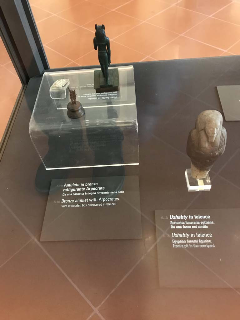 VIII.7.28 Pompeii. April 2019. Bronze amulet with Harpocrates, on left, which was found in a wooden box in the cella.
Egyptian funeral figurine found in a pit in the courtyard, on right.
Now in Naples Archaeological Museum. Photo courtesy of Rick Bauer.