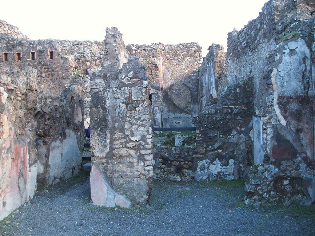 IX.2.7 Pompeii. December 2004. Looking east to rear room of shop (b) with corridor to rear (d) on north side of it.
According to Minervini –
From the shop-room, by a threshold of white marble, one entered into another small room.
The flooring was of opus signinum with a rough mosaic of white stones in the centre.
The walls were white, with panels of different colours, decorated with masks, birds and animals: then a drum suspended by ribbons, a small half-open basket, and another uncertain object. From here and there, on the two side walls, you could see a bearded figure, with wide tunic, who held a vase in the shape of a kantharos on his head with both hands. In the wall on the right are shown two Cupids, partly faded, and as many on the wall to the left, one with patera the other with lyre, and all with fluttering cloak: many times Cupid with lyre appeared in pompeian paintings.
At the top of the wall, there are residues of stucco composed of a flat fascia with pretty foliage, and a curve with palm leaves and stalks (caulicles): in the space comprised by these two lines a hippocampus and two dolphins are painted.
See Bullettino Archeologico Napoletano Nuova Serie: Anno Primo, no.4, Agosto 1852, p.25. Minervini.
