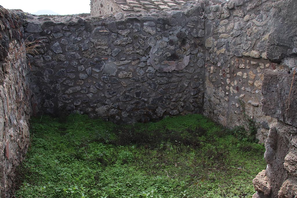 IX.7.19 Pompeii. October 2024. Room k, looking south through doorway in corridor f. Photo courtesy of Klaus Heese.
The east wall and south-east corner are on the left, presumably the site of the lararium painting, but nothing now remains.
According to Boyce, a small room, room k, located between kitchen and garden on the south side of the house, was originally part of the garden.
This small room had a doorway into the kitchen.
Then the room was separated from the garden and the doorway to the kitchen was bricked-up.
On the east wall of this new room was a lararium, with a Genius standing to the right of a tripod.
On the other side of the tripod stood the tibicen. Below and to the right, ran a camillus.
Below this were two serpents, gliding, one from each side, amongst plants towards an altar.
Between this painting and the south-east corner of the room were painted, two pots, a large bottle, sausages, a calf’s head, ribs of pork on a spit, and a phallus.
He references – Not. Scavi, 1880, 491; Bull. Inst., 1883, 82.
See Boyce G. K., 1937. Corpus of the Lararia of Pompeii. Rome: MAAR 14. (p.88, no.438)
According to NdS, the were also two Lares:
To right in the atrium there is a room communicating with the back room of the following taberna, and the access to the kitchen with the latrine, and to a cell, where the lararium painting is divided into two strips; in the lower part there are the two serpents, which rush towards the altar with the offerings, and in the upper part the sacrificing Genius Familiaris, with tibicen and a camillus between the two Lares.
See Notizie degli Scavi di Antichità, 1880, p. 491.
Fröhlich notes: Boyce does not mention Lares.
Boyce 438 (IX 7, 19) describes the Genius with Camillus and Tibicen grouped around the altar in the usual way, but without the Lares.
See Fröhlich, T., 1991. Lararien und Fassadenbilder in den Vesuvstädten. Mainz: von Zabern. (p.112).
See Giacobello, F., 2008. Larari Pompeiani: Iconografia e culto dei Lari in ambito domestico. Milano: LED Edizioni, (p.209, no.106)