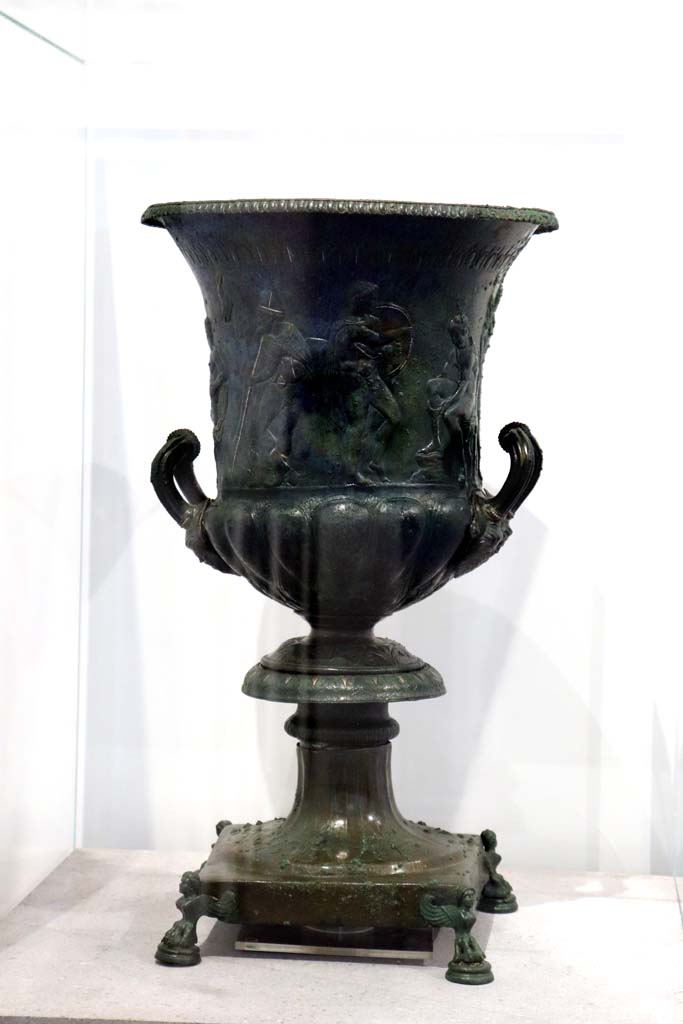 IX.13.1-3, Pompeii. February 2021.
Bronze krater with scenes in relief, found in triclinium, on display in Antiquarium, VIII.1.4.
Photo courtesy of Fabien Bièvre-Perrin (CC BY-NC-SA).
According to the description card in the Antiquarium -
The bronze krater was decorated with eight armed male figures, six standing and two sitting.
One of the seated figures is tying his shoelaces.
A young man stands in front of him holding a shield, with a bearded figure holding a staff behind.
Young and naked, the second seated figure caresses an animal with one hand and holds a sword by the hilt in the other.