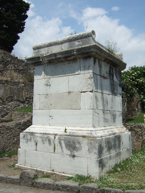 HGE37 Pompeii. May 2006. Marble plaque on east side. On the plaque is the inscription:
M. ALLEIO LVCCIO LIBELLAE PATRI 
AEDILI II VIR PRAEFECTO QVINQ(uennali) ET 
M. ALLEIO LIBELLAE F DECVRIONI VIXIT 
ANNIS XVII LOCVS MONVMENTI
PVBLICE DATVS EST ALLEIA M F 
DECIMILLA SACERDOS PVBLICA 
CERERIS FACIVNDVM CVRAVIT VIRO 
ET FILO

This differs from the south side plaque where the last word is FILIO rather than FILO.
The plaque is also narrower and the inscription is on more (eight) lines.
According to Epigraphik-Datenbank Clauss/Slaby (See www.manfredclauss.de) this reads

M(arco) Alleio Luccio Libellae patri
aedili IIvir(o) praefecto quinq(uennali) et 
M(arco) Alleio Libellae f(ilio) decurioni vixit 
annis XVII locus monumenti
publice datus est Alleia M(arci) f(ilia)
Decimilla sacerdos publica
Cereris faciundum curavit viro
et filio      [CIL X 1036 second part]

Gell describes this as “A well-executed tomb, in stone, of simple form, about 15 feet high. 
Upon two of its sides are similar inscriptions, which inform us that it was erected by Alicia Decimilla, public priestess of Ceres, to her husband Luccius Libella, aedile, duumvir, and quinquennial prefect : also to her son M. Alleius Libella, decurion at 17, upon ground decreed by the public for that purpose.”
See Gell, W, and Gandy J. P., 1819.  Pompeiana. London: Rodwell and Martin.  (p. 111).