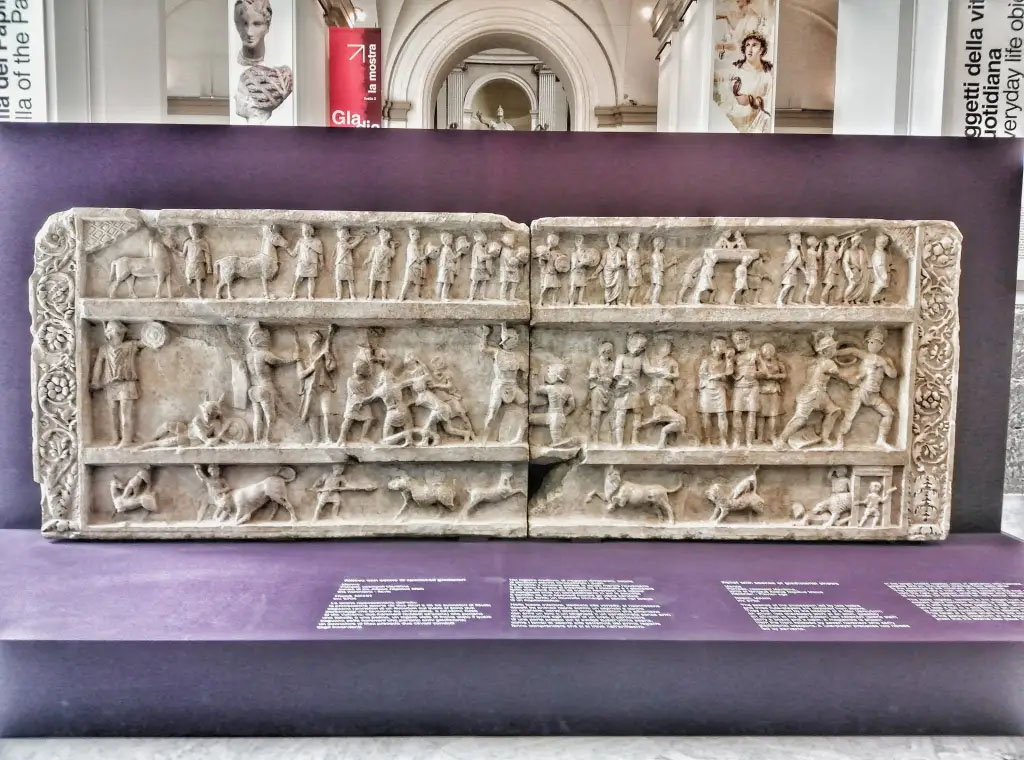 SG6 Pompeii. 2021. Gladiatorial relief with hunting scenes from a tomb at Porta Stabia. The background gives an indication of its large size.
According to Emmerson, this relief measures more than 4 metres long and 1.5 metres high.
See Emmerson A., 2010. Reconstructing the Funerary Landscape at Pompeii's Porta Stabia: Rivista di Studi Pompeiani 21, pp. 78, fig. 1.
According to Osanna, in view of what remains and the visible traces, as well as archive research that is still ongoing, it is more than likely that the top of the tomb SG6, damaged by the construction of the San Paolino building in the nineteenth century, was completed with the gladiator relief preserved in MANN. If you evaluate the role that gladiatorial shows and venations have in praise, it can be assumed that the well-known relief with gladiatorial scenes and hunts with animals kept in Mann was placed there.
The relief has dimensions compatible with the monument SG6, as its length is about 4 metres long, and would respond well in the theme to the role of the deceased as an extraordinary organizer of games. 
Moreover, the relief, discovered by the superintendent Avellino in the 1840s, was found out of place (from the damage evidently suffered by the monument in the construction of San Paolino), precisely in the area of Porta Stabia.
Now in Naples archaeological Museum. Inventory number 6704.

SG6 Pompeii. 2021. Rilievo gladiatore con scene di caccia da una tomba a Porta Stabia. Lo sfondo dà un'indicazione delle sue grandi dimensioni.
Secondo Emmerson, questo rilievo misura oltre 4 metri di lunghezza e 1,5 metri di altezza.
Vedi Emmerson A., 2010. Reconstructing the Funerary Landscape at Pompeii's Porta Stabia: Rivista di Studi Pompeiani 21, pp. 78, fig. 1.
Secondo Osanna, in considerazione di quanto resta e delle tracce visibili, oltre che delle ricerche di archivio tuttora in corso, è più che verosimile che la parte superiore della tomba SG6, danneggiata dalla costruzione dell’edificio di San Paolino nell’Ottocento, fosse completata con un rilievo conservato al MANN. Se si valuta il ruolo che gli spettacoli gladiatori e le venationes hanno nell’elogio, si può ipotizzare che fosse ivi collocato il noto rilievo con scene gladiatorie e di caccie con animali conservato al Mann. 
Il rilievo ha infatti dimensioni compatibili con il monumento SG6, lungo com’è circa 4 m., e risponderebbe bene nel tema al ruolo del defunto di straordinario organizzatore di giochi. 
Inoltre, il rilievo, scoperto dal soprintendente Avellino negli anni ’40 del XIX secolo, fu rinvenuto fuori posto (per i danni subiti dal monumento evidentemente per la costruzione di san Paolino), proprio nell’area di porta Stabia.
Ora al Museo archeologico di Napoli. Numero di inventario 6704.

