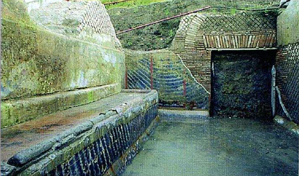 Complesso dei triclini in località Moregine a Pompei. Excavation photo. The kitchen with the large bench and the doorway leading to the triclinium area.
The kitchen (F) is large, 4.40 x 7.05 meters wide with the long and wide cooking bench against the eastern wall (4.75 x 1.40 metres); a large door to the south allowed one to reach the triclinium area, while another door to the north, on the west wall, allowed one to reach the uncovered service area (fig. 5). The cocciopesto floor has a slope towards the north-east corner of the room, here, just at the corner, a sort of pillar contains a large gutter made of terracotta tubules (about 22 cm in diameter) which probably collected part of the roof water. The decoration of the walls with a black background is very simple as is characteristic of rustic environments as well. the lararium scene in the centre of the west wall: all the walls are divided into panels delimited by red bands, alternately decorated with simple white streaks now arched now oblique. Likewise, the walls of the cooking counter. At the centre of the west wall is a large lararium painted with scenes of sacrifice on a white background. In the foreground, in the centre, we see an altar to which two long snakes approaching, rich in details as regards the scales. In the background, also in the centre, there is a priest, head veiled, dressed in a white mantle with red borders. On the sides two attendants with ivy crowned heads and short white robes act as helpers.
See Nappo, S. C., 2005. I triclini di Murecine uso ed interpretazione, p. 59, fig. 5.